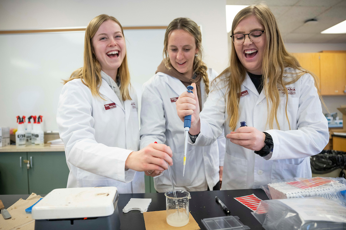 Three students in white lab coats share a laugh 